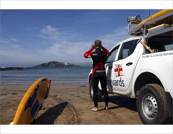 A lifeguard on Bantham beach monitoring the sea through a pair of binoculars. The lifeguard is stood next to a 4x4 patrol vehicle and yellow rescue board