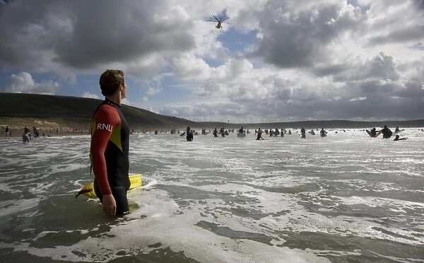 An RNLI lifeguard in the sea at Woolacombe beach, Devon. Lots of people in the water swimming and surfing, helicopter overhead