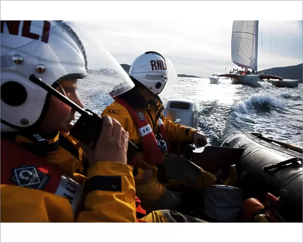 Two crew members from Barra RNLI on an ILB in the foreground, one talking into a radio. Yacht competing in the Round Britain and Ireland yacht race 2010 in the background. Leisure