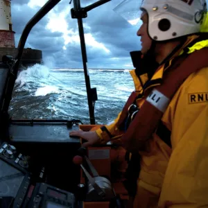 Coxswain stood at the helm of the Berwick-upon-Tweed Mersey class lifeboat Joy and Charles Beeby 12-32, lots of spray, lighthouse in the background