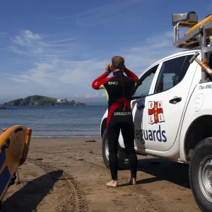 A lifeguard on Bantham beach monitoring the sea through a pair of binoculars. The lifeguard is stood next to a 4x4 patrol vehicle and yellow rescue board