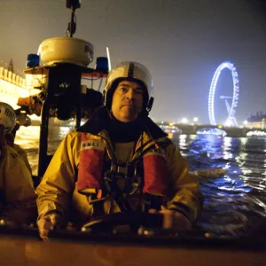 Tower lifeboat crew members on board an E-class lifeboat on the River Thames at night, Houses of Parliament and the London Eye in the background