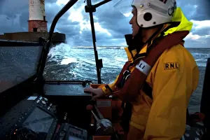 Coxswain stood at the helm of the Berwick-upon-Tweed Mersey class lifeboat Joy and Charles Beeby 12-32, lots of spray, lighthouse in the background